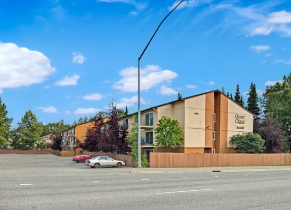 Three-story apartment building with "Quiet Creek" sign, surrounded by trees, under a blue sky with clouds. Two cars parked in front on a paved lot.