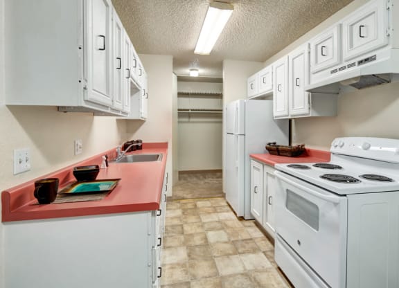 Compact kitchen here at Quiet Creek Apartments with a retro feel features white cabinets, a red countertop, and white appliances. Bright lighting highlights the clean, orderly space.