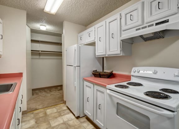 Narrow kitchen here at Quiet Creek Apartments with white cabinets, red countertops, and a white stove. A white refrigerator stands beside an open pantry with empty shelves. Bright lighting.