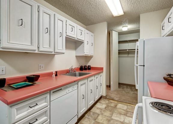 Narrow kitchen here at Quiet Creek Apartments with coral countertops, white cabinets, stove, and fridge. Bright lighting, clean, organized, with a modern yet cozy feel.