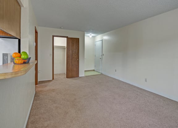 Empty living room here at Rancho Tudor Apartments with beige carpet and white walls, featuring a wooden door to a closet, a white entrance door, and a bar counter with a fruit bowl.