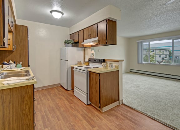 Cozy kitchen here at Rancho Tudor Apartments with wooden cabinets, white appliances, and a laminate floor opens to a carpeted living area with a large window, offering natural light.