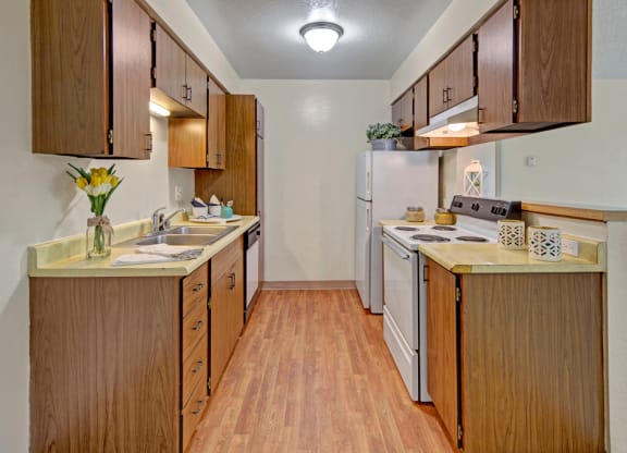 A narrow kitchen here at Rancho Tudor Apartments with wood cabinets and laminate flooring. Yellow flowers in a vase are on the counter, alongside neatly arranged kitchenware, creating a warm, welcoming ambiance.
