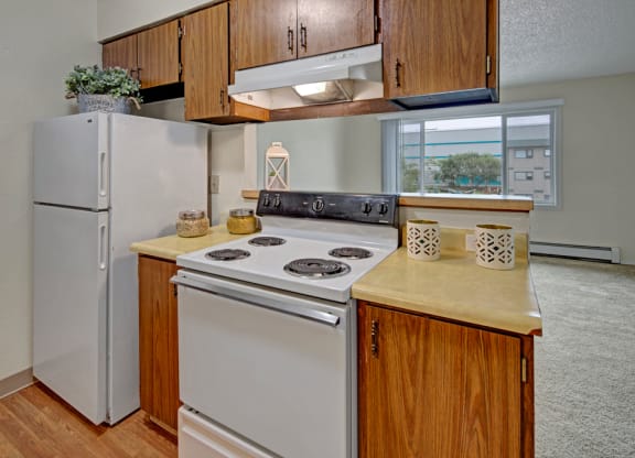 Compact kitchen here at Rancho Tudor Apartments with wood cabinets, white appliances including a stove and fridge. Decor includes plants and lanterns, with ample natural light. Cozy ambiance.