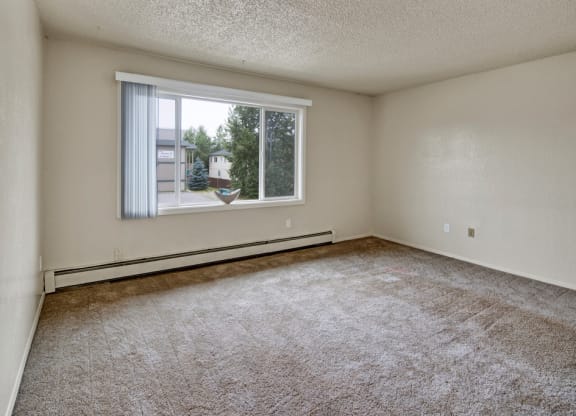 An empty beige room here at Rancho Tudor Apartments with a large window showing trees and buildings outside. The carpet is brown, the walls are plain, and there's a minimal, neutral feel.