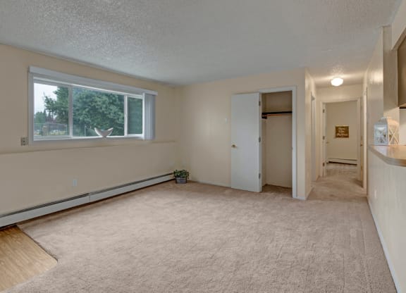 Empty beige living room here at Rancho Tudor Apartments with a large window showing greenery outside. The room features carpet, an open closet with a door, and a hallway with dim lighting. Calm and neutral ambiance.