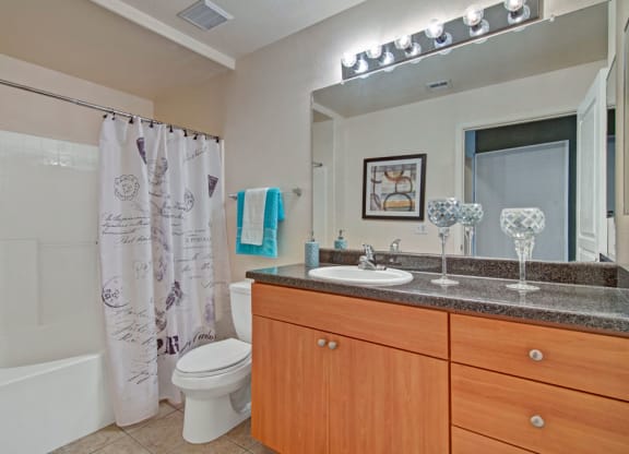 Modern bathroom here at Rio Paradiso Apartments with wooden vanity and granite countertop. A large mirror with bright lights, decorative candles, and a patterned shower curtain add elegance.