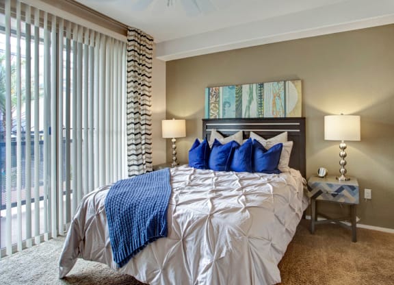 A cozy bedroom features a neatly made bed with a cream comforter and blue pillows here at Rio Paradiso Apartments. Two lamps on wooden nightstands illuminate the space. Vertical blinds cover a large window, creating a serene atmosphere.