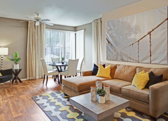 Modern living room here at Rio Paradiso Apartments with beige sectional sofa, yellow and black cushions, abstract rug, and dining area. Large rope bridge photo on wall, adding warmth.