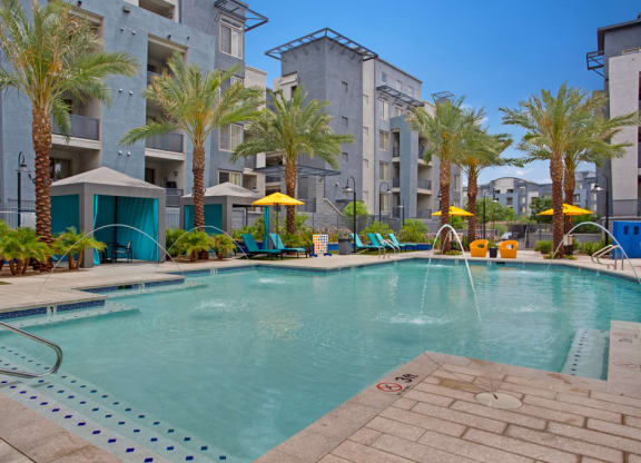 Rio Paradiso Apartment complex pool area with palm trees and blue-gray buildings. Features lounge chairs, yellow umbrellas, cabanas, and water fountains, creating a relaxing atmosphere.