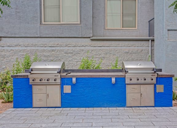 Outdoor BBQ area here at Rio Paradiso Apartments with two stainless steel grills on a vibrant blue counter. The background features a textured gray wall and greenery, creating a welcoming space.