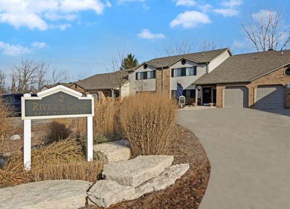 Sign reading "River's Cove Apartment Homes" near a pathway leading to brick and siding apartments with garages under a clear blue sky.