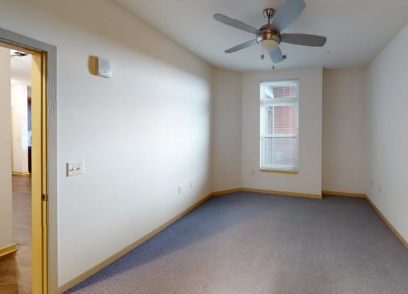 An empty room with beige walls and gray carpeting features a ceiling fan and a window with blinds, creating a neutral and spacious atmosphere at Royster Commons Apartments.