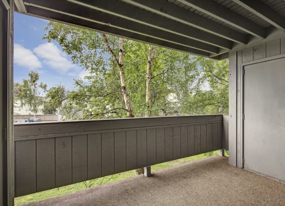 Covered balcony here at Russian Jack Apartments with wooden railing, beige carpet, and closed gray door, overlooking lush green trees. Bright sky and distant buildings create a peaceful setting.