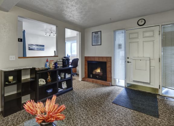 Cozy office entrance here at Russian Jack Apartments with a lit fireplace, shelf with snacks and supplies, and a large clock above a white door flanked by blinds. Warm ambiance.