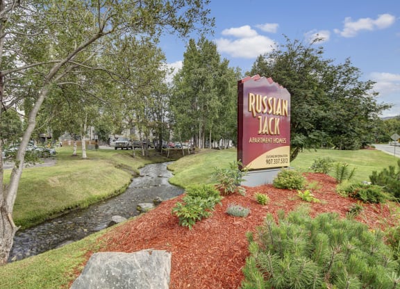 Sign for Russian Jack Apartment Homes by a small creek with red mulch landscaping, surrounded by lush trees and a clear blue sky, creating a peaceful setting.