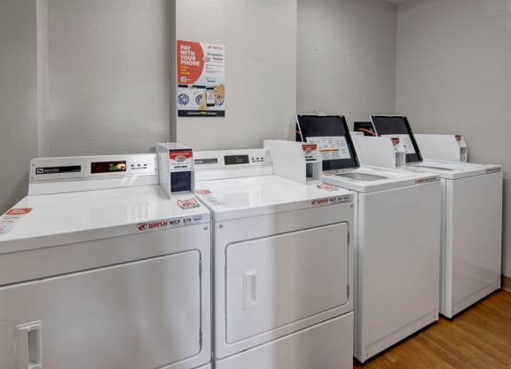 A row of white commercial washers and dryers is in a clean, gray laundry room here at Russian Jack Apartments. A poster on the wall advertises mobile payment options.