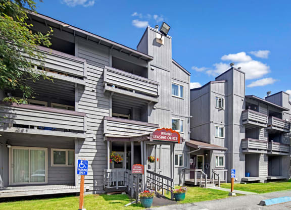 Gray, multi-story Russian Jack Apartment building with balconies under a blue sky. A leasing office sign is visible. Accessible parking signs are in front.