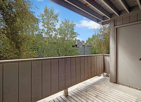 A wooden balcony here at Russian Jack Apartments with railing offers a view of trees and distant buildings. A closed door is on the right, under a partially covered roof, in daylight.