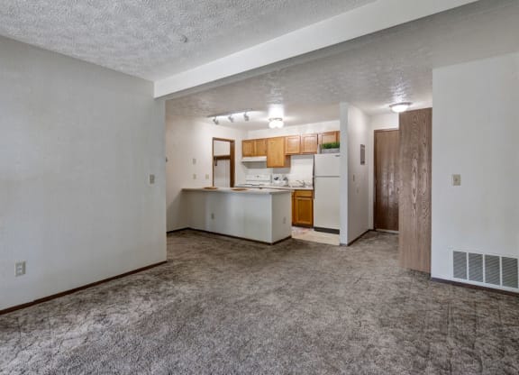 Empty living room here at Russian Jack Apartments with beige carpet, white walls, and an adjacent kitchen featuring wooden cabinets, white appliances, and a small island. Cozy and neutral.