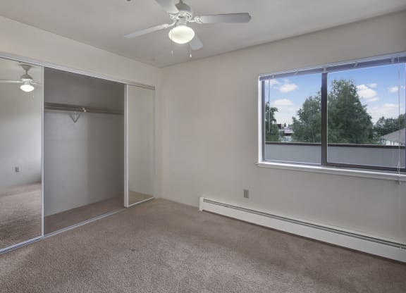 Empty bedroom here at Silver Ridge Apartments with beige carpet and white walls. Large mirrored closet doors reflect a ceiling fan. Bright window view reveals green trees and blue sky.