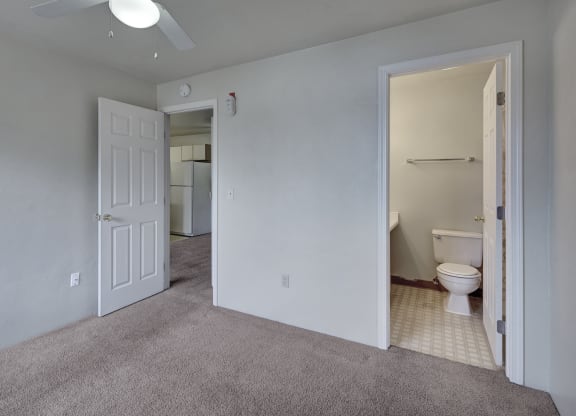 Empty bedroom here at Silver Ridge Apartments with gray walls and carpet, open door to a bathroom with a toilet and towel rack. Ceiling fan overhead. Doorway reveals a kitchen area with a fridge.