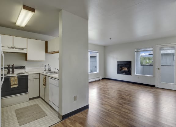 A small kitchen here at Silver Ridge Apartments with light cabinets, white appliances, and a cozy rug opens into a bright living room with hardwood floors and a corner fireplace.