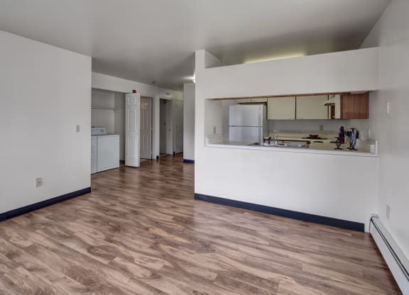 Spacious, empty living room here at SIlver Ridge Apartments with light wood flooring and white walls. Open kitchen with a visible fridge and countertop, providing a clean, minimalist feel.