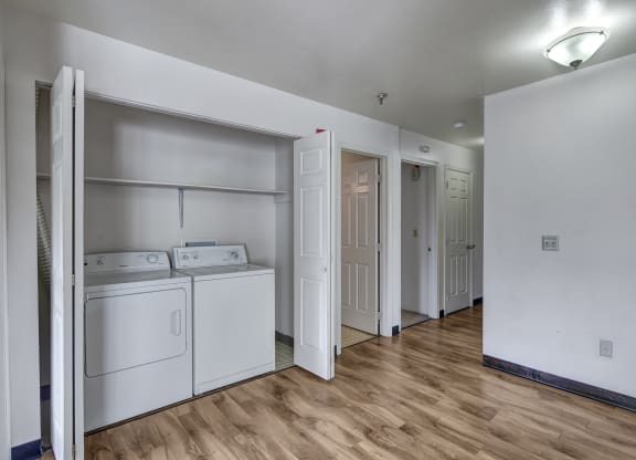 Bright laundry area here at Silver Ridge Apartments with white washer and dryer set against a light wall. Wood flooring leads to open doors revealing more rooms, under soft overhead lighting.