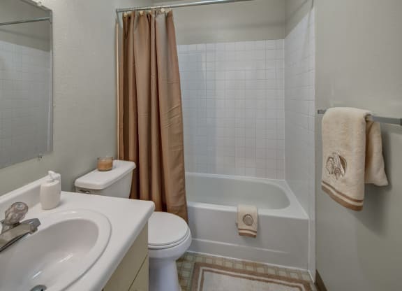 A small bathroom here at Silver Ridge Apartments with white sink and toilet, beige shower curtain, and matching towels with shell motifs. A candle adds a warm, inviting touch.