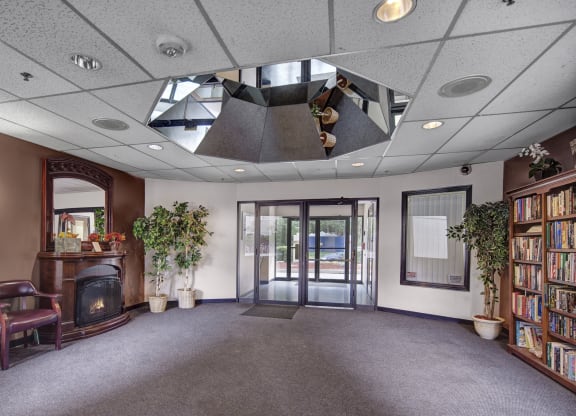 Lobby here at Silver Ridge Apartments with a cozy and inviting feel, featuring a dark wooden fireplace with a mirror above it, two potted plants, a bookshelf full of books, and seating.