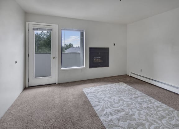Empty living room here at Silver Ridge Apartments with white walls, a glass door, and a window showing trees outside. A fireplace is set into the wall, and a carpet with floral patterns lies on the floor.