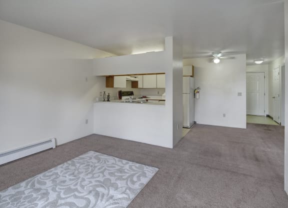 Spacious living room here at Silver Ridge Apartments with beige carpet and patterned rug. Open view of a white kitchen with appliances. Ceiling fan and soft lighting create a cozy atmosphere.