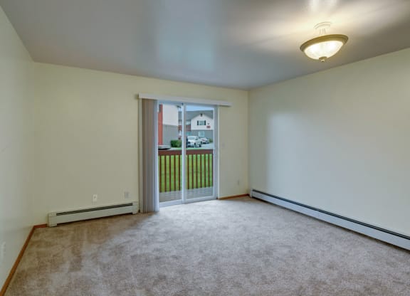 Empty beige room here at Stawberry Lane Apartments with carpet, bright ceiling light, and sliding glass doors leading to a small balcony overlooking grass and residential houses. Calm ambiance.