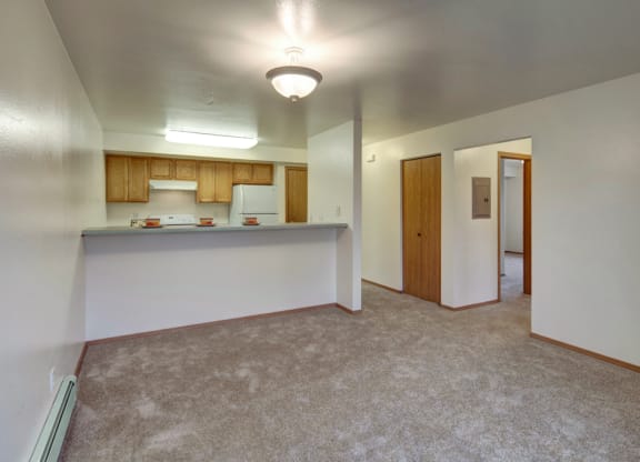 Open-plan apartment interior here at Strawberry Lane Apartments with beige carpet, white walls, and ceiling light. Kitchen features wooden cabinets, stove, and fridge. Doorway leads to another room.