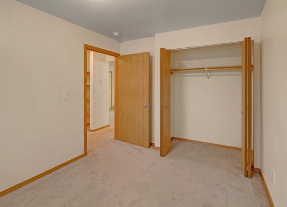 Empty beige room here at Strawberry Lane Apartments with an open closet on the right and a wooden door on the left. Soft carpet flooring, simple and clean atmosphere.