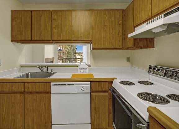 Compact kitchen here at Susitna Ridge Apartments with wooden cabinets, white appliances, and a stainless steel sink. A window view of trees and buildings adds warmth to the space.