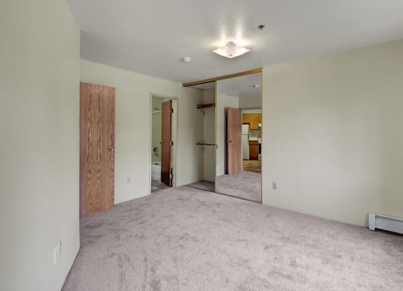 Empty room here at Susitna Ridge Apartments with beige carpet, mirrored closet doors reflecting a kitchen, wooden doors leading to a bathroom; minimalistic and bright atmosphere.