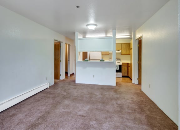 Spacious, empty apartment living room here at Susitna Ridge Apartments with beige carpet and white walls, a hallway with wooden doors, and a small kitchen with light wood cabinets.