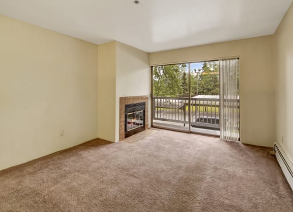 Empty living room here at Susitna Ridge Apartments with beige carpet and walls, featuring a tiled fireplace. A large sliding glass door leads to a balcony with views of greenery. Bright and serene atmosphere.