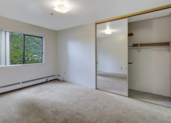 Empty, bright bedroom here at Susitna Ridge Apartments with beige carpet, large window showing greenery, and mirrored sliding closet doors reflecting ceiling light, creating a serene atmosphere.