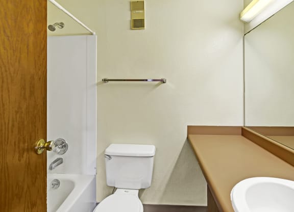 Compact bathroom here at Susitna Ridge Apartments with a white tub, shower, and toilet. Brown countertop with a sink and large mirror. Beige walls create a neutral, minimal tone.