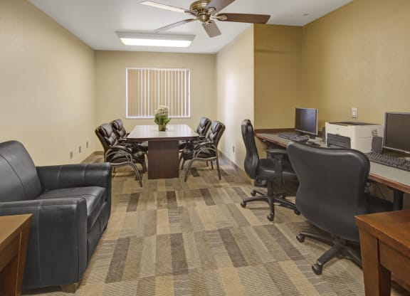 Susitna Ridge Apartment's modern, neutral-toned office features a round table with six leather chairs, a black sofa, two computers on desks, and a window with blinds.