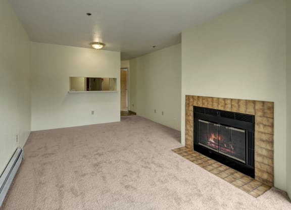 Empty living room here at Susitna Ridge Apartments with beige carpet, featuring a lit fireplace with a brown tile surround on the right, and a small serving hatch on the left wall. Cozy ambiance.