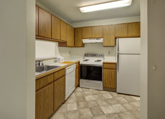 Compact kitchen here at Susitna Ridge Apartments with wooden cabinets, white appliances including a stove, refrigerator, dishwasher, and a sink. Tiled floor and bright lighting.