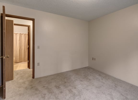 Empty beige carpeted bedroom here at Taiga Apartments with a doorway to a bathroom, partially visible beige shower curtain. Neutral tones convey a calm, minimalist atmosphere.