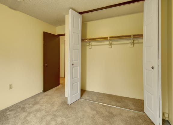 Empty beige room here at Taiga Apartments with open closet doors, revealing a wooden rod and shelf inside. Carpeted floor and an open brown door leading to another room. Quiet and clean atmosphere.