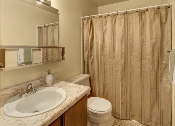 A small bathroom here at Taiga Apartments with a marble-patterned sink, soap dispenser, and toothbrush holder. Beige shower curtain, wooden door with towels, bright lighting.