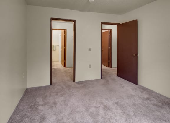 Empty bedroom here at Taiga Apartments with beige carpet, white walls, and open brown doors leading to two additional rooms. The atmosphere is neutral and minimalistic.