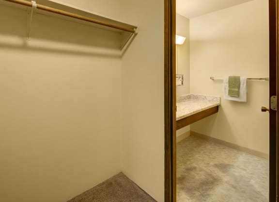 Minimalist interior here at Taiga Apartments showing an empty closet with a wooden rod and shelf. Adjacent bathroom features a marble countertop and towel rack with towels.
